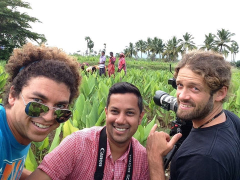 Three smiling men, one with a video camera, in a leafy crop field with farm workers and palm trees behind