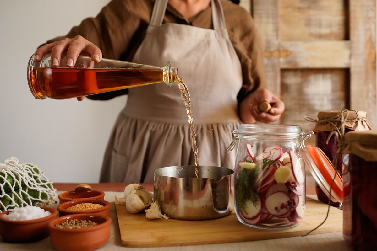 A cook pours amber vinegar from an unlabelled bottle into a pot beside a jar of sliced radishes, garlic and dill
