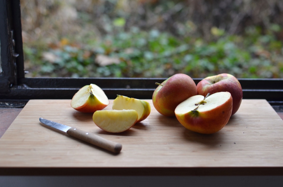 Halved and sliced red-and-yellow apples with a paring knife on a wooden board by a window