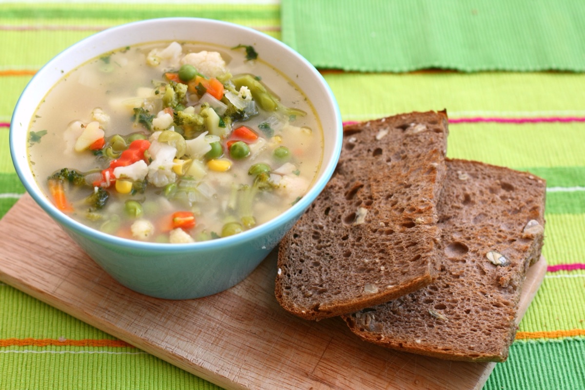 A bowl of vegetable soup with peas, carrots and cauliflower beside two slices of brown bread