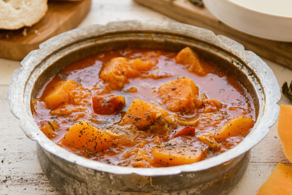 A metal pot of stew with chunks of orange squash in a tomato broth, sliced squash beside it
