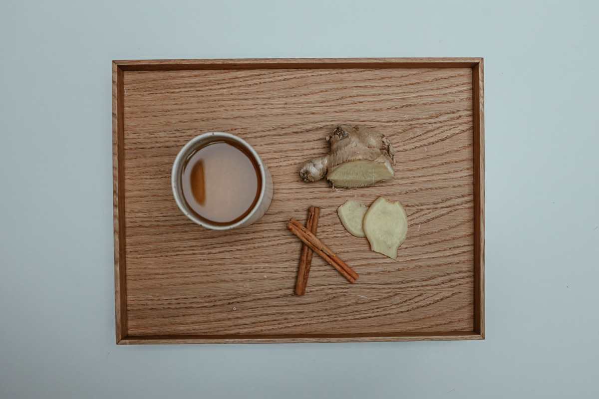 A cup of tea beside fresh ginger root, ginger slices and two cinnamon sticks on a wooden tray