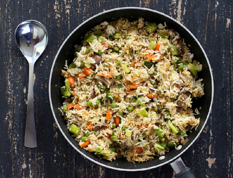 Skillet of rice cooked with carrots, broccoli, peas and mushrooms beside a spoon