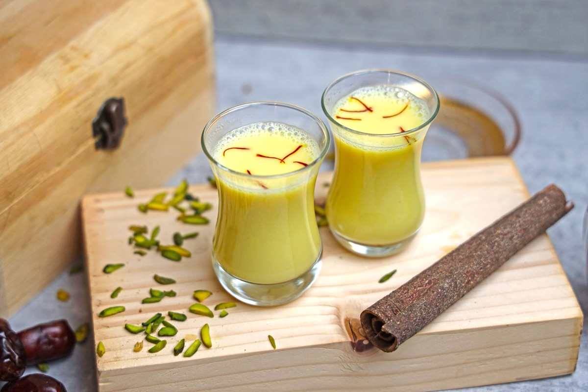 Two small glasses of turmeric and saffron milk topped with saffron threads on a wooden board with pistachios and cinnamon