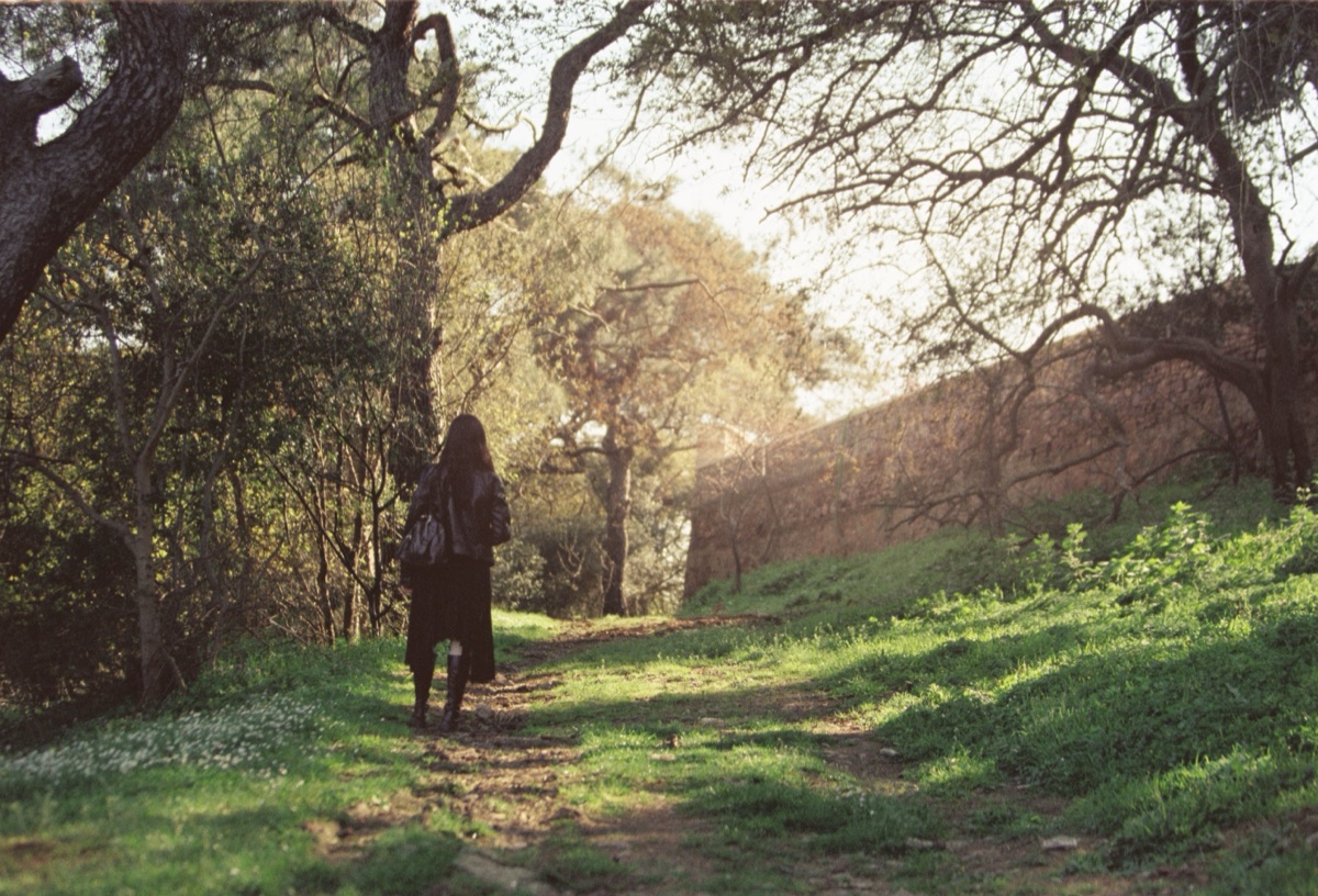A woman seen from behind walks alone along a grassy path under trees in low golden light