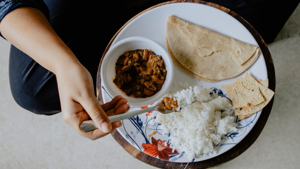 Seen from above, a seated person eats a plate of rice, rajma, roti and papad with a fork