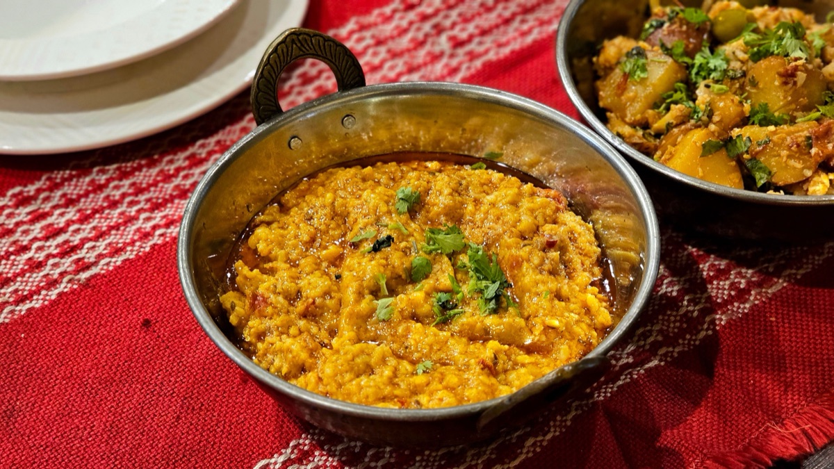 A metal pan of thick yellow lentils topped with cilantro beside a bowl of spiced potatoes on a red woven tablecloth