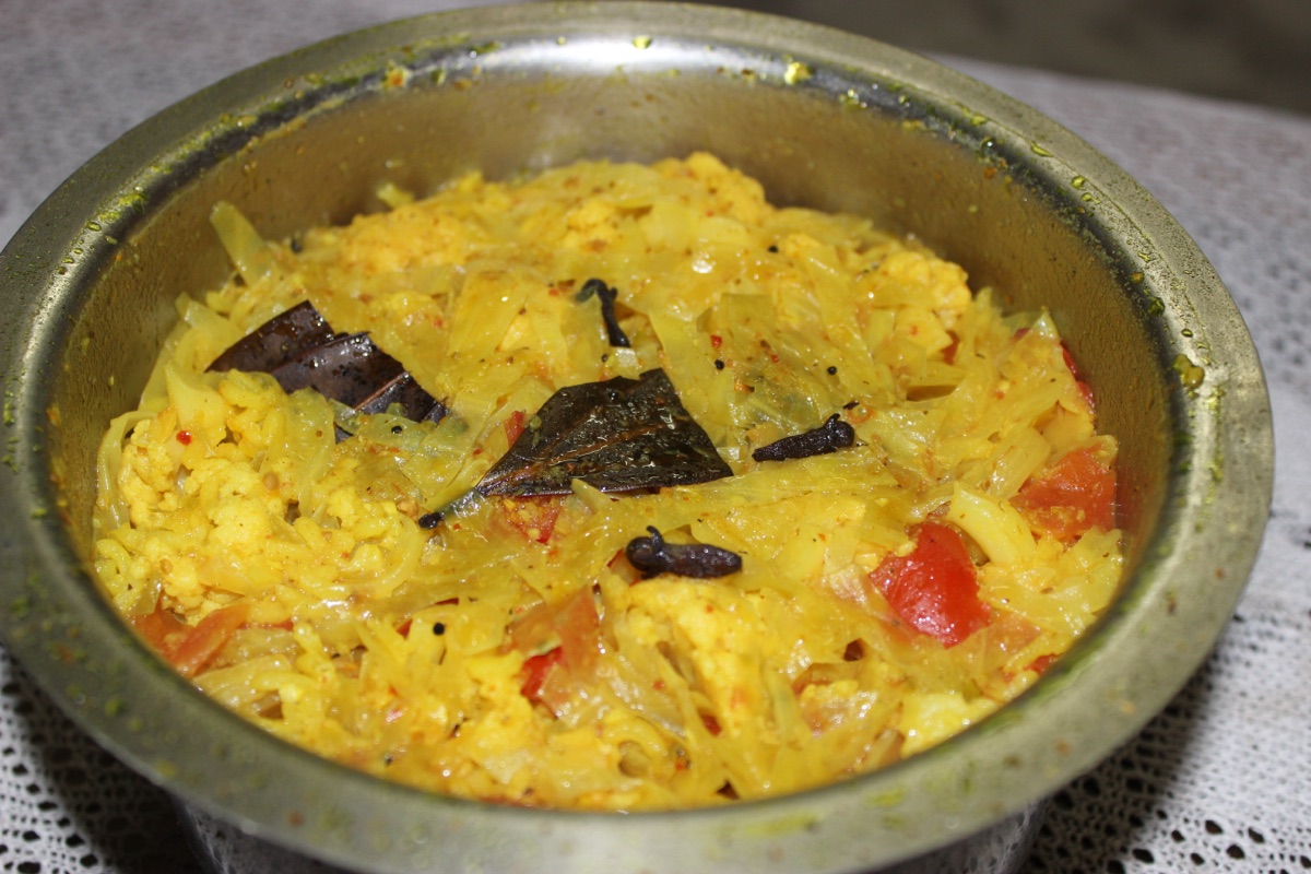 A steel bowl of yellow Gujarati masala khichdi with cabbage, tomato, bay leaves and cloves