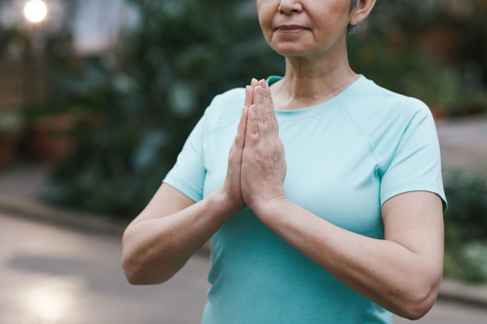 A grey-haired woman in a mint T-shirt holds her palms together in front of her chest, plants blurred behind her