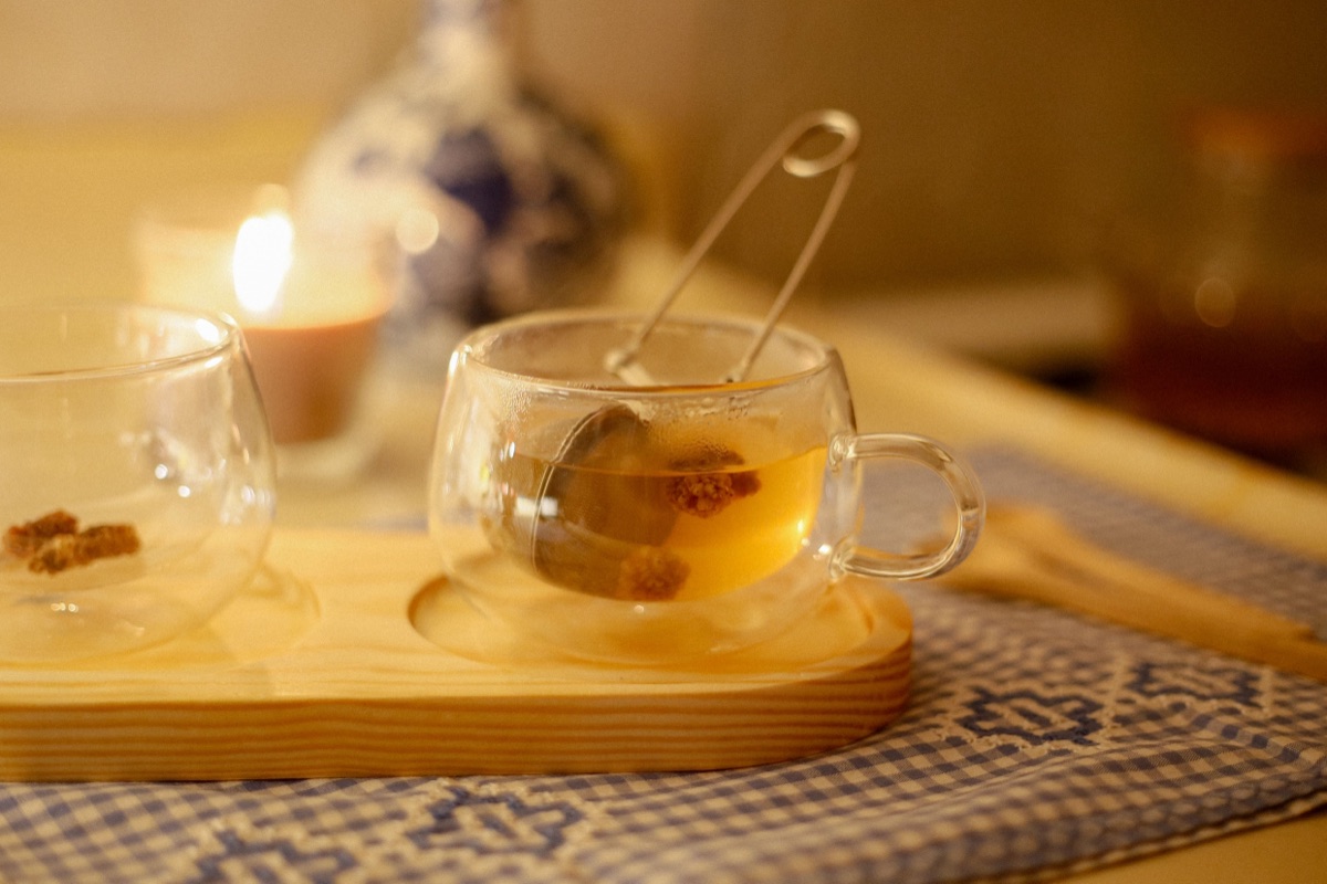 A glass cup of tea with a steel infuser on a wooden tray, a lit candle glowing behind it