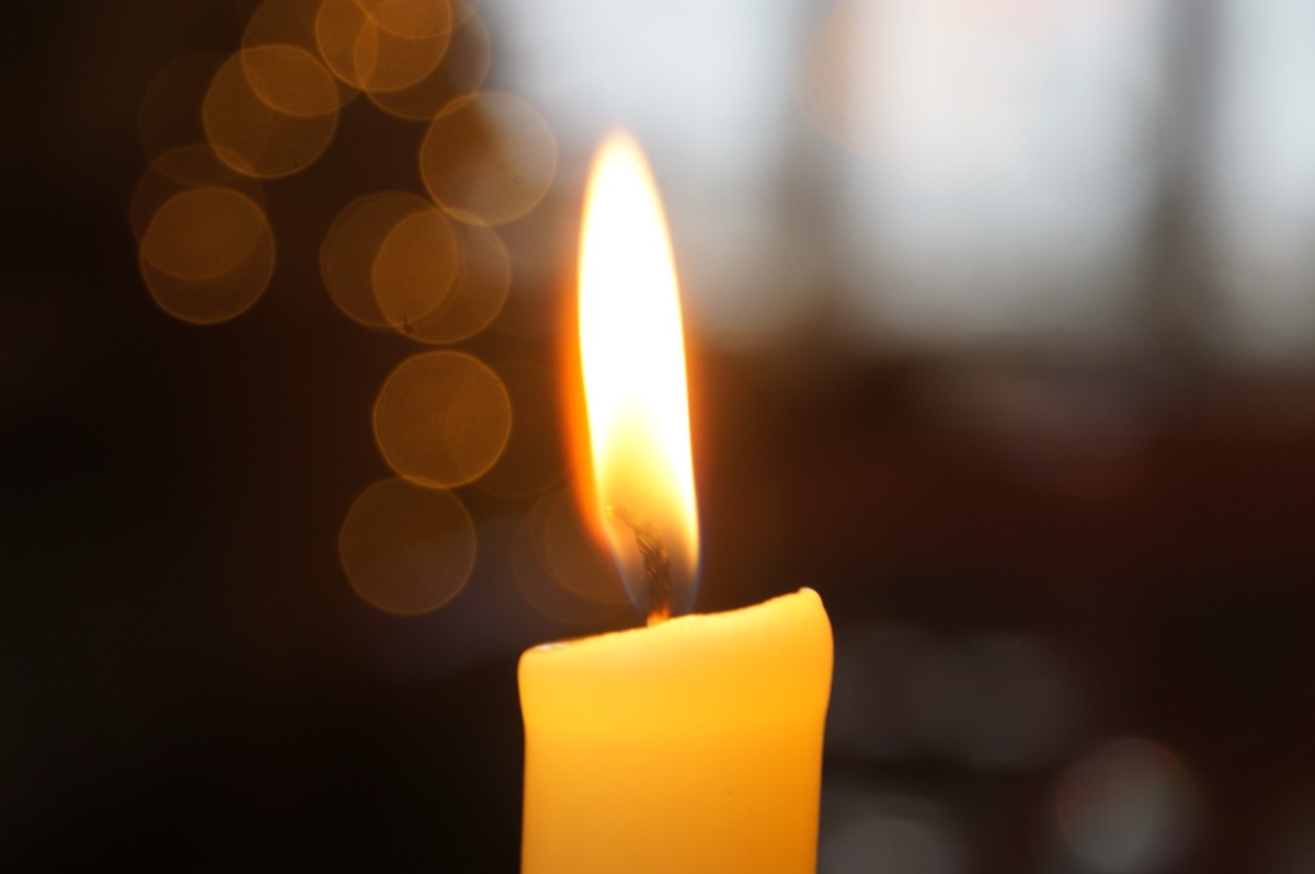 A single yellow candle burning with a tall, steady flame in a dim, softly blurred room