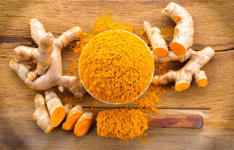 Fresh turmeric roots around a bowl of ground turmeric powder with a wooden spoon