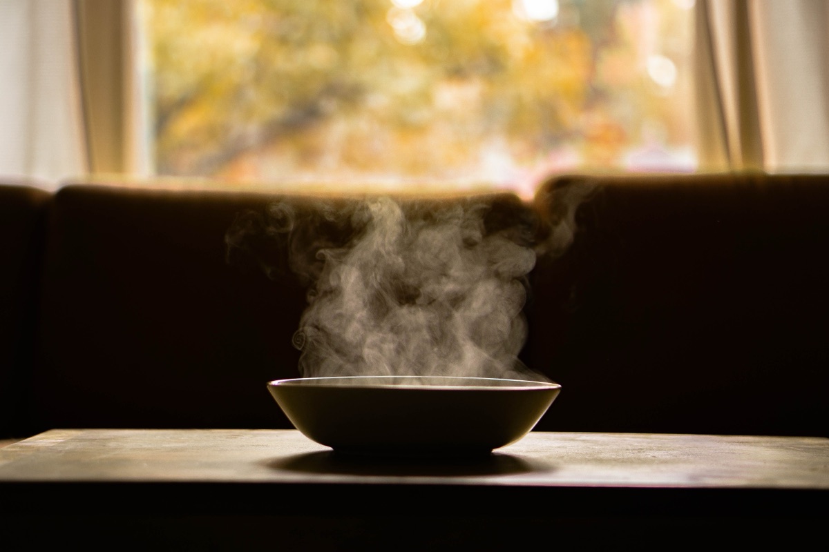 A steaming bowl of soup on a dark wooden table with soft window light behind it