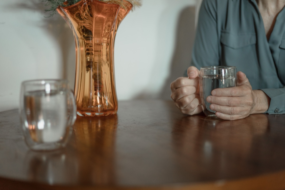 Hands holding a glass mug of water on a round wooden table beside an amber glass vase