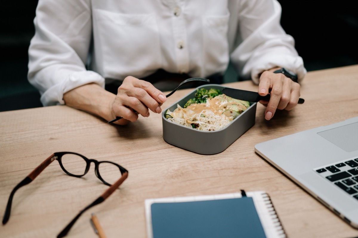 Hands with a fork and knife over noodles and greens in a lunch box on a desk, beside a laptop and glasses