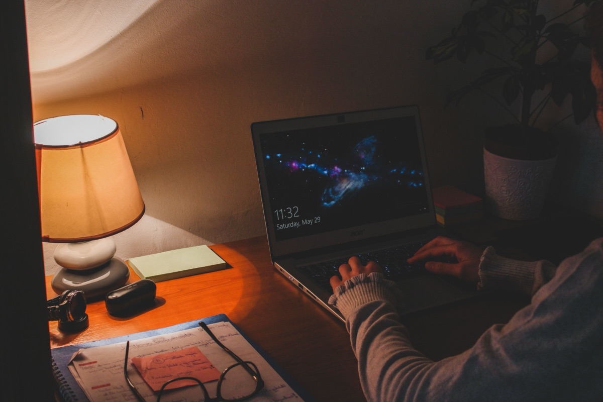 Hands typing on a laptop at night beside a lit table lamp, a notepad and a pair of glasses