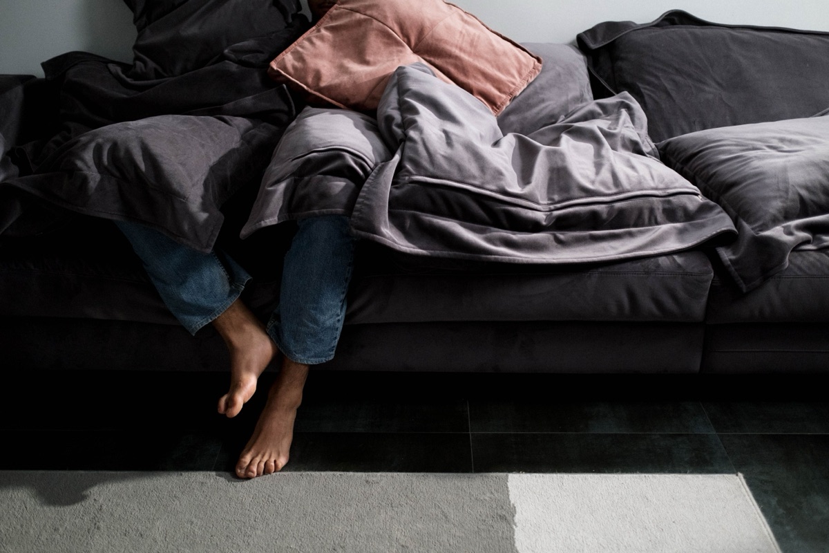 A person hidden under grey blankets and a pink cushion on a dark sofa, only jeans and bare feet showing