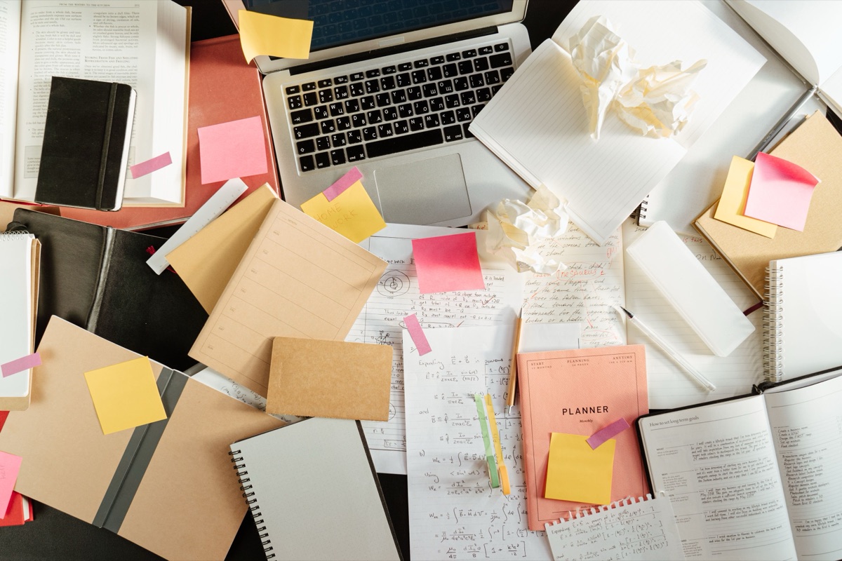 A cluttered desk of open notebooks, sticky notes and crumpled paper around a laptop, seen from above