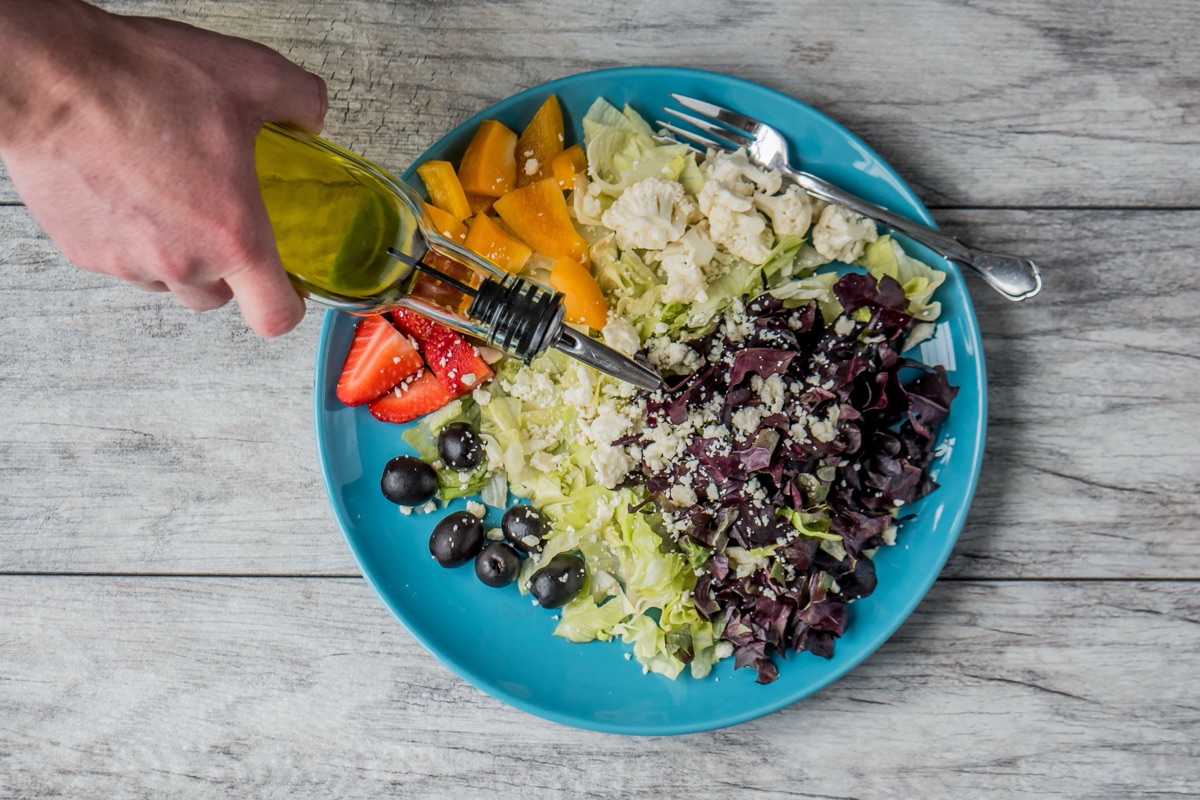 Hand pouring oil from a bottle over a salad of lettuce, red lettuce, cauliflower, orange pepper, olives, strawberries and crumbled cheese
