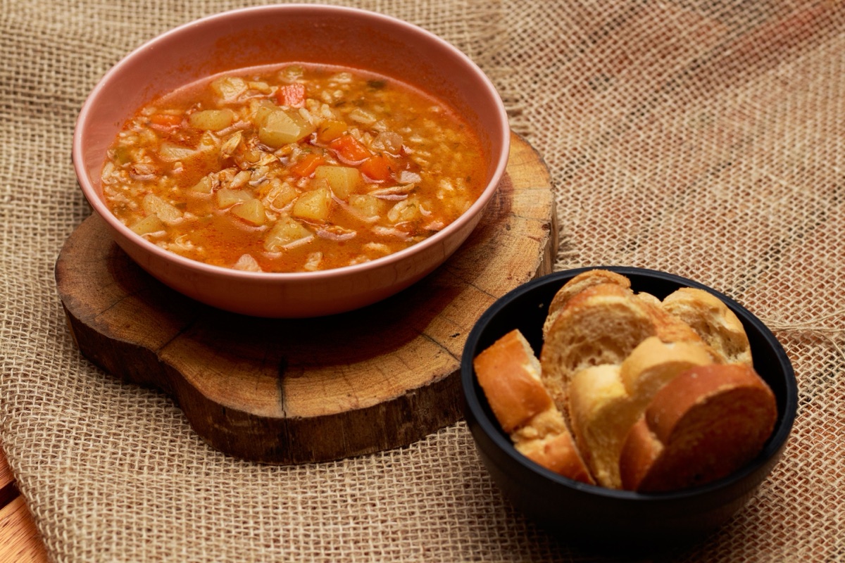 Bowl of vegetable soup on a wooden board beside a dark bowl of sliced bread on burlap