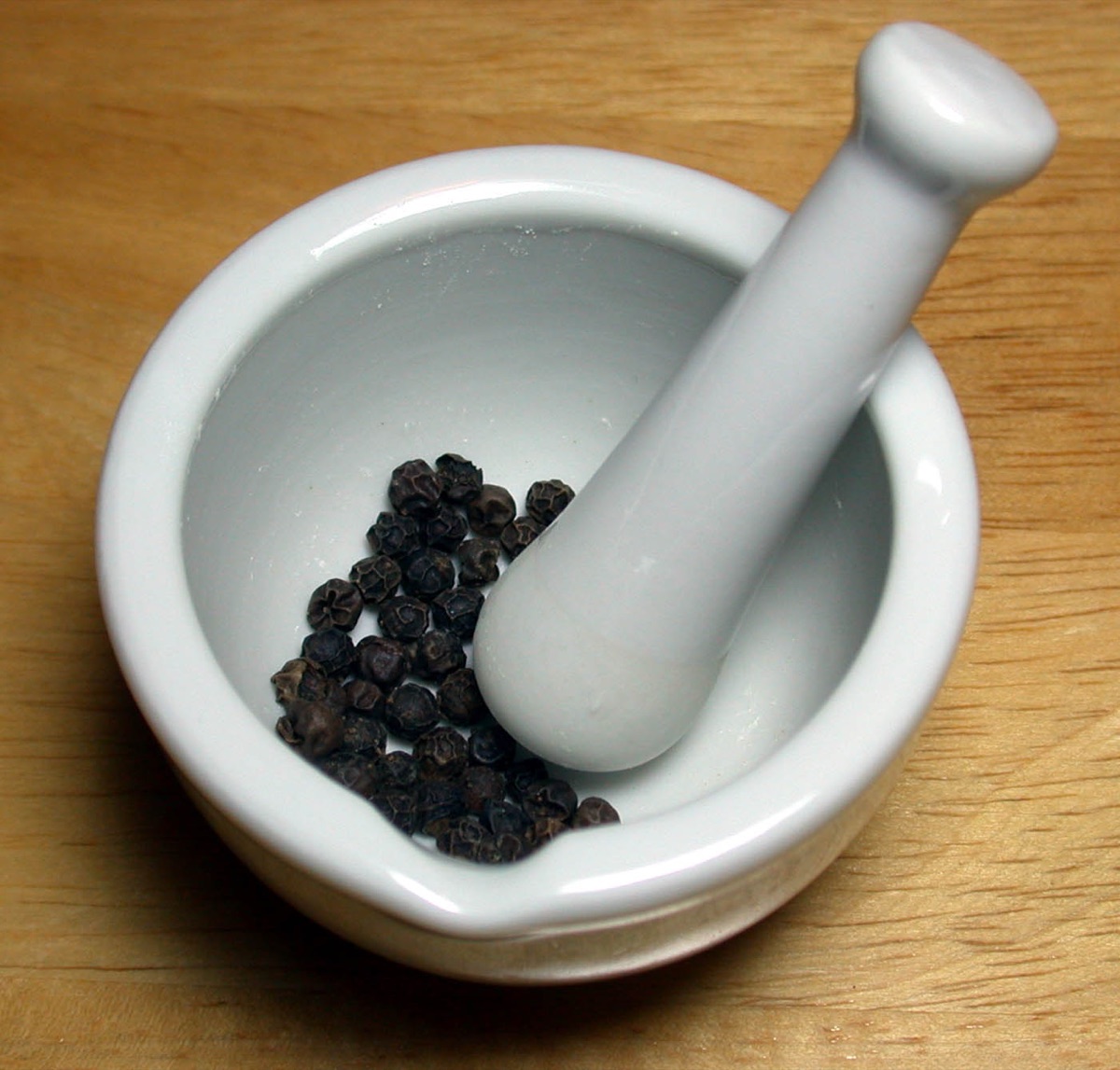 Whole black peppercorns in a white ceramic mortar with its pestle, on a wooden counter