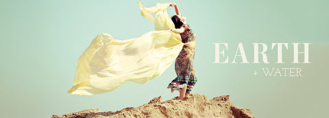 Woman on a rocky peak holding a flowing pale yellow scarf in the wind, with the words Earth + Water
