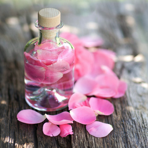 Corked glass bottle of rose water filled with pink rose petals, more petals scattered on wood