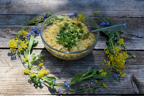 Glass bowl of yellow quinoa and mung bean kitchari topped with fresh herbs, ringed by wildflowers