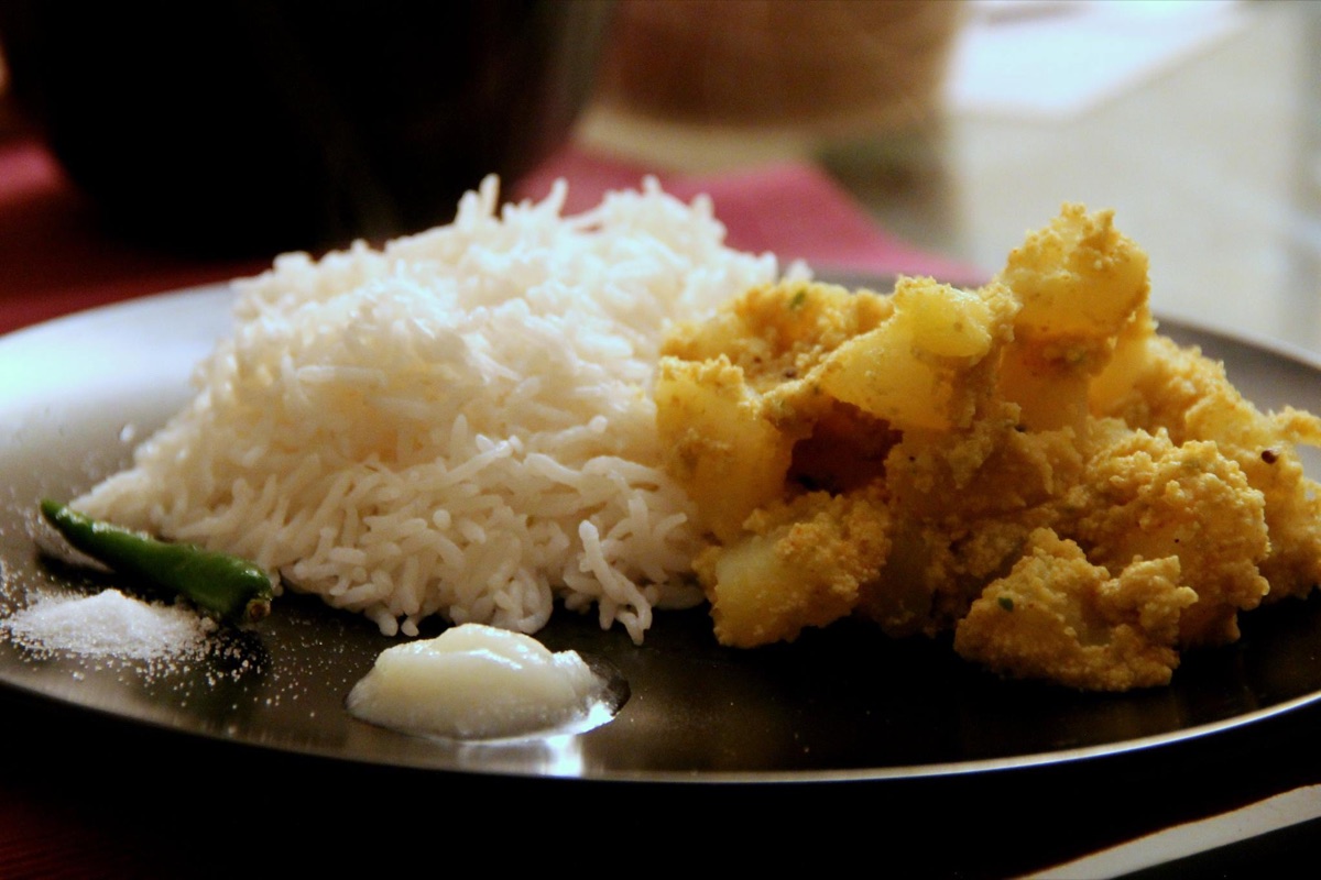 Potato pieces coated in pale poppy seed paste beside a mound of white rice and a green chilli on a dark plate