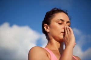 Woman with eyes closed practicing alternate nostril breathing, fingers at her nose, against a blue sky