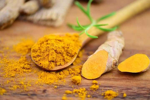Wooden spoon of ground turmeric powder beside sliced fresh turmeric root on a wooden table