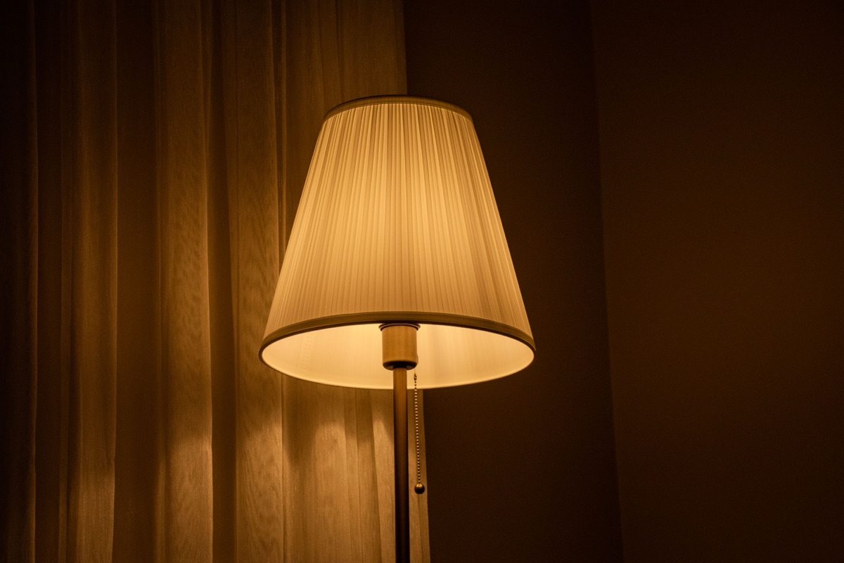 A pleated fabric lampshade glowing warm beside a curtain in a dark room