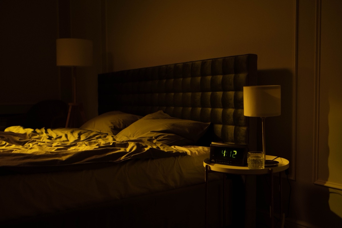 A dim bedroom with an unmade bed and a bedside digital clock reading 4:12