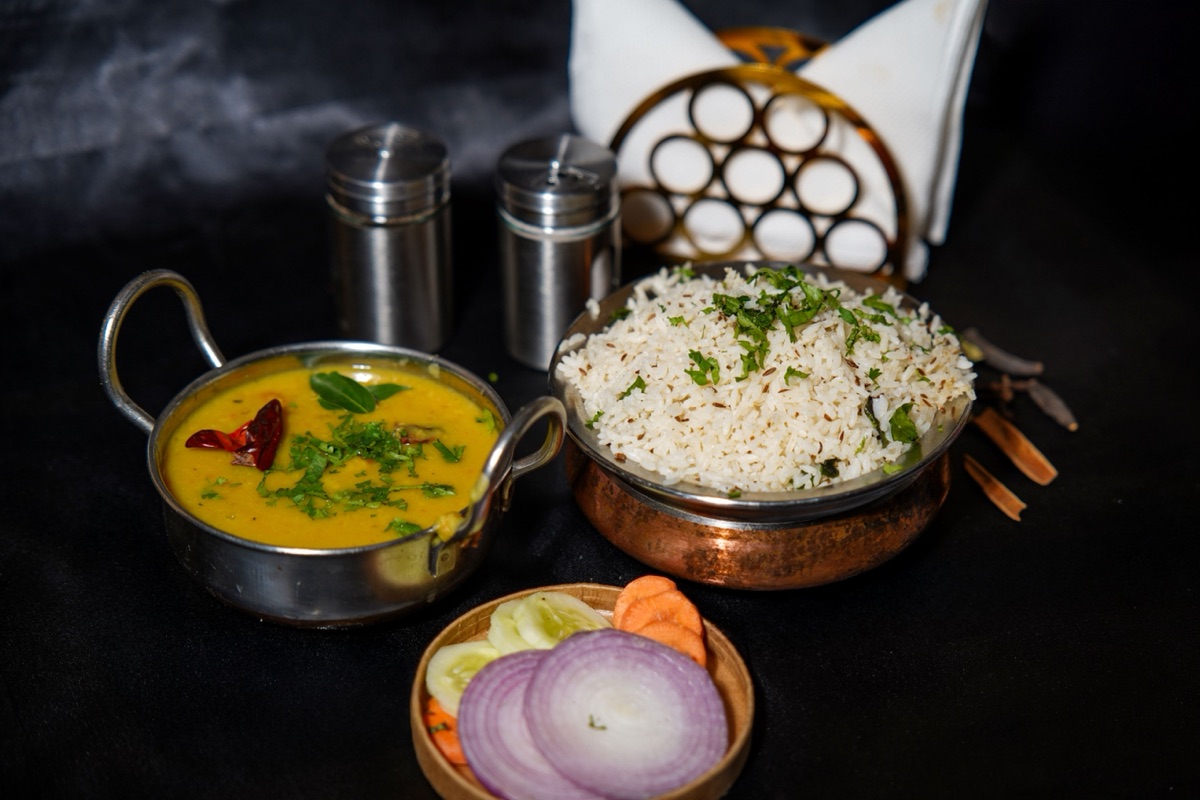 A steel pot of yellow dal tadka with dried red chilli beside a bowl of jeera rice