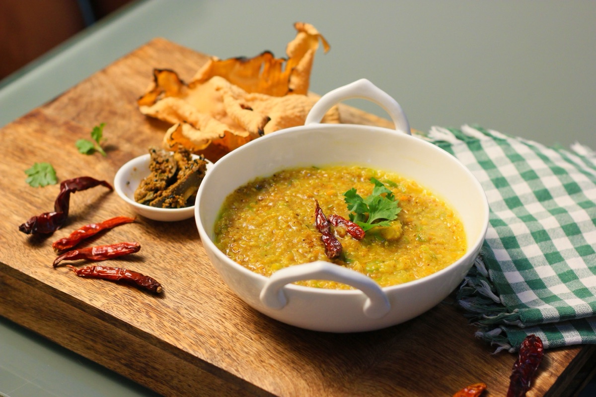 A white bowl of yellow khichdi topped with coriander and dried red chilies, with papad and pickle on a wooden board