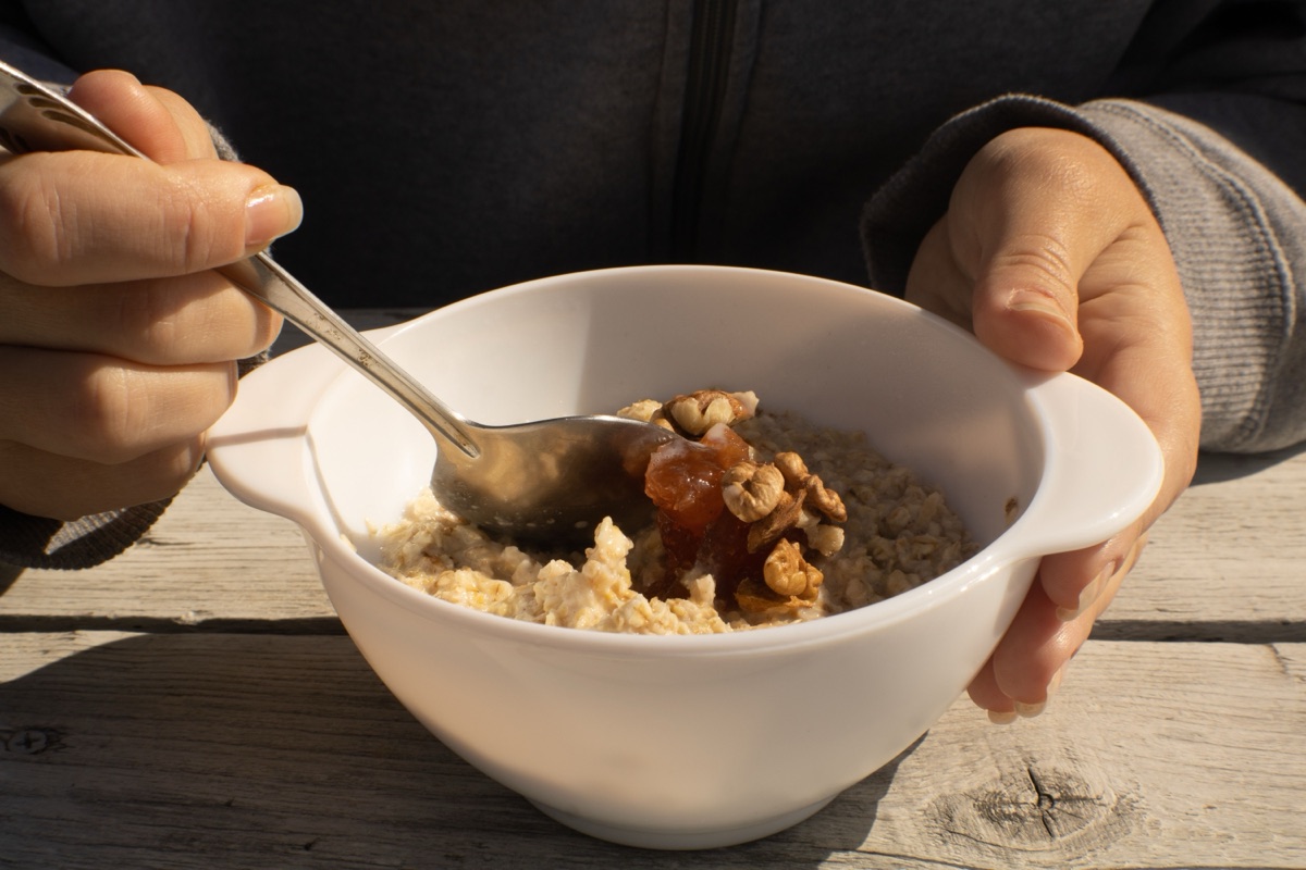 One hand holds a white bowl of cooked oatmeal topped with walnuts and jam while the other dips a spoon in, over a wooden table