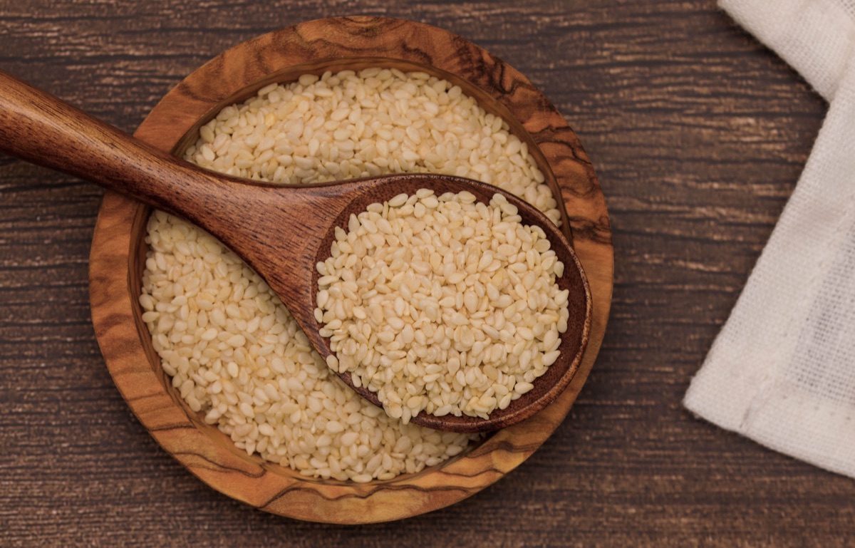 A round olive-wood bowl and a wooden spoon full of pale hulled sesame seeds on a dark wooden table