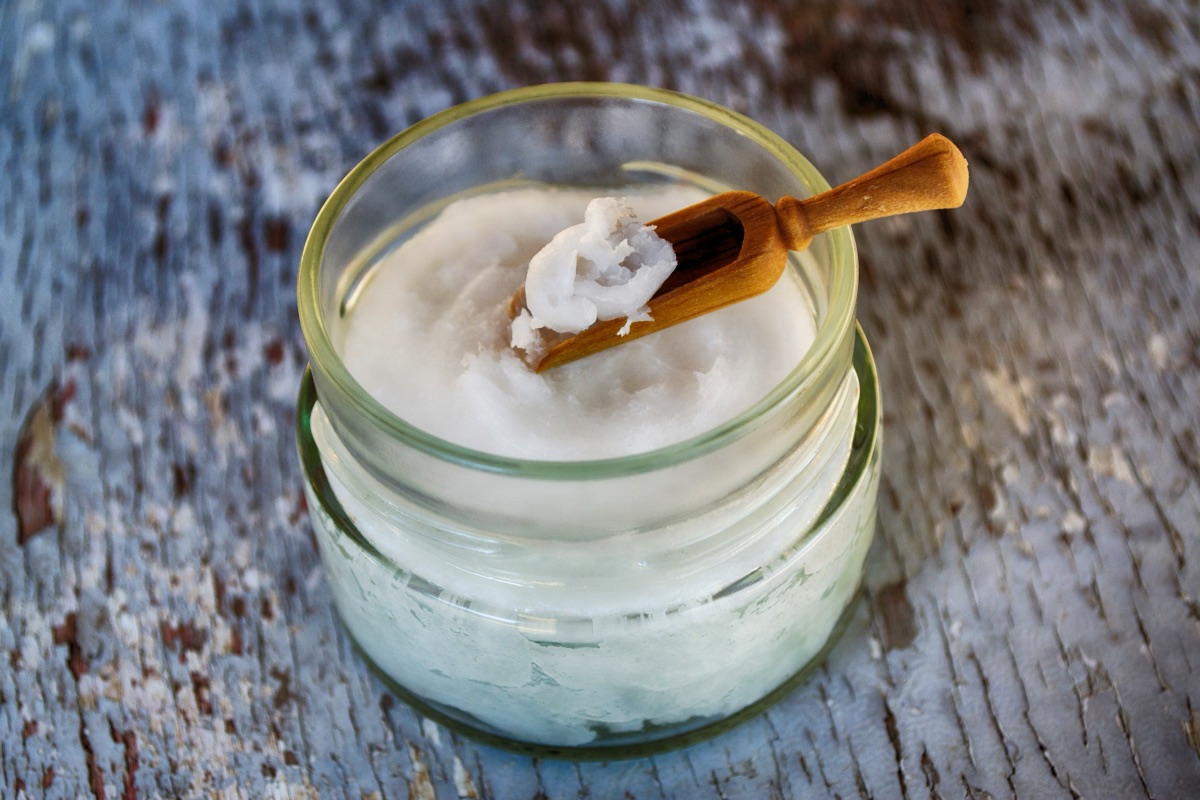 A glass jar of solid white coconut oil with a small wooden scoop resting on top, on weathered painted wood