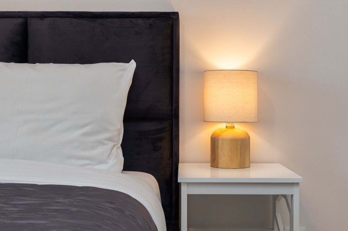 A lit table lamp with a wooden base and linen shade on a white nightstand beside a bed with a white pillow
