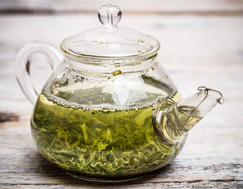 Glass teapot of brewing green tea with loose tea leaves on a weathered wooden table