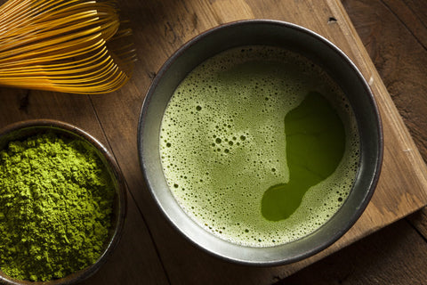 Bowl of frothy whisked matcha green tea beside a bamboo whisk and a dish of matcha powder