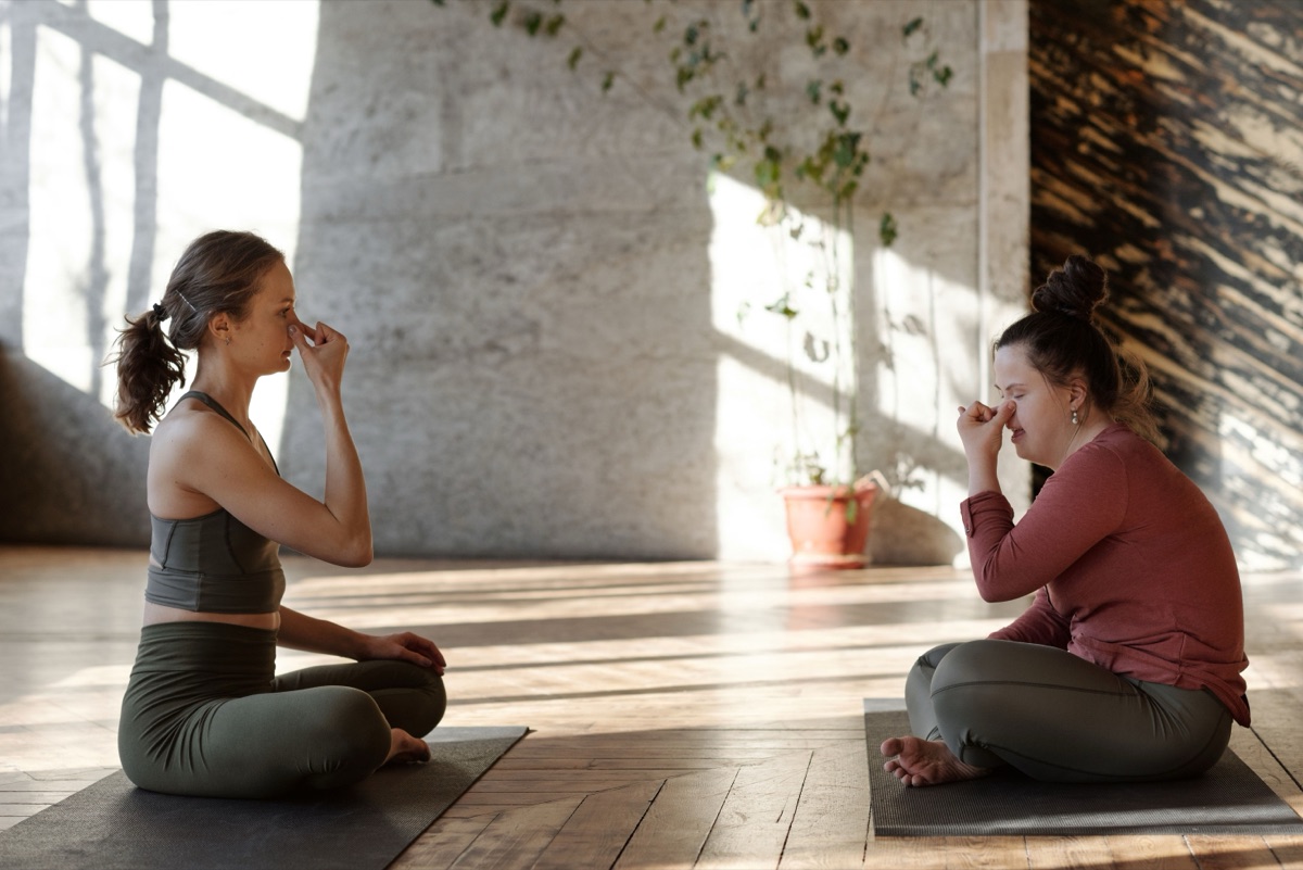 Two women seated cross-legged on mats in a sunlit studio, each with a hand raised to her nose, eyes closed