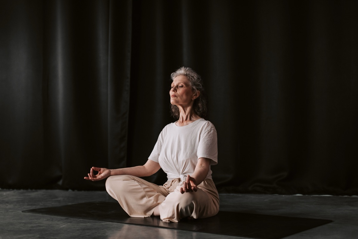 A grey-haired woman in a white T-shirt sits cross-legged on a mat against a dark curtain, eyes closed, hands resting by her knees