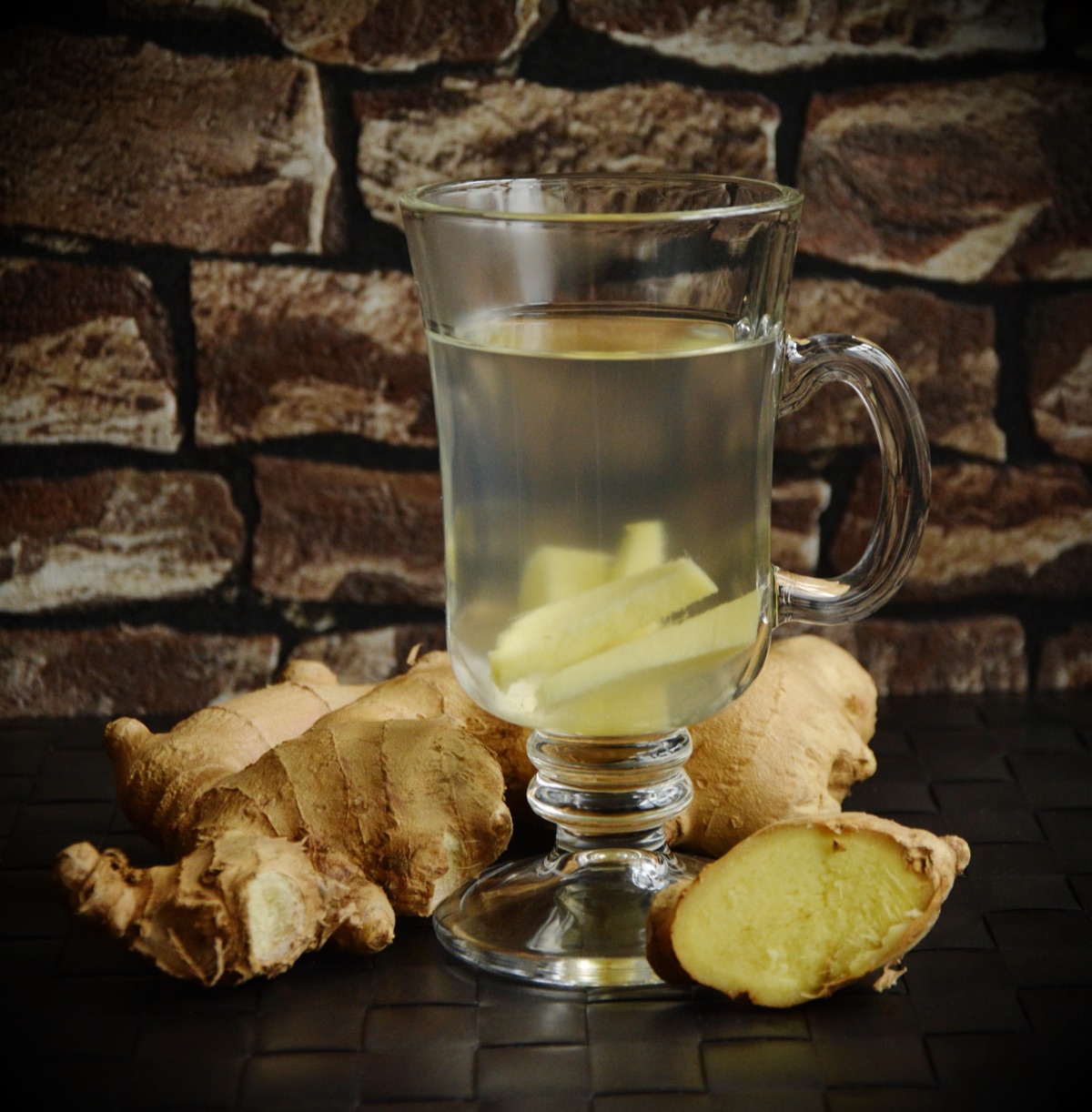 A glass mug of pale ginger tea with ginger slices inside, beside whole fresh ginger roots