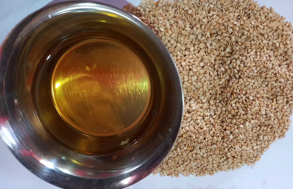 A steel bowl of golden sesame oil beside a heap of sesame seeds on a white surface