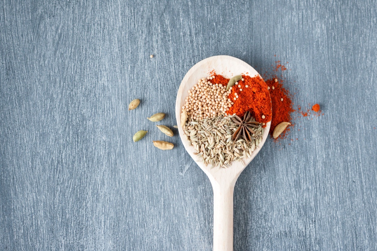A wooden spoon holding yellow mustard seeds, fennel seeds, red chili powder and a star anise, with cardamom pods beside it
