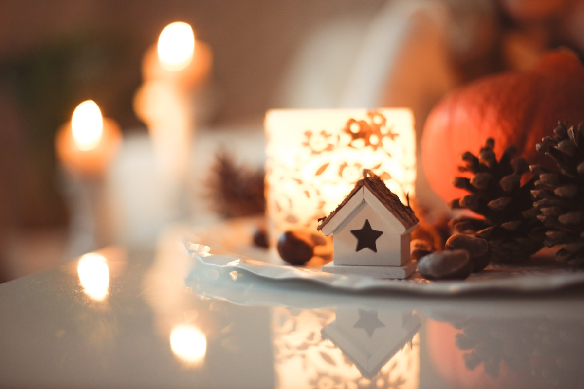 Lit candles and a glowing patterned candle lantern beside pine cones, chestnuts, a pumpkin and a tiny wooden house ornament