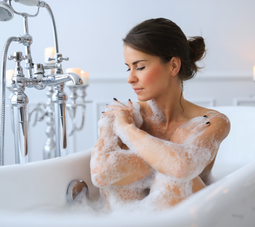 Woman in a white bathtub covering her arms and shoulders with bath foam