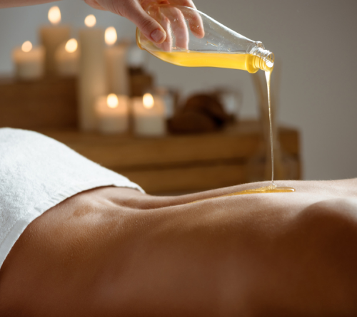 Warm golden massage oil poured from a bottle onto a person's lower back, candles behind