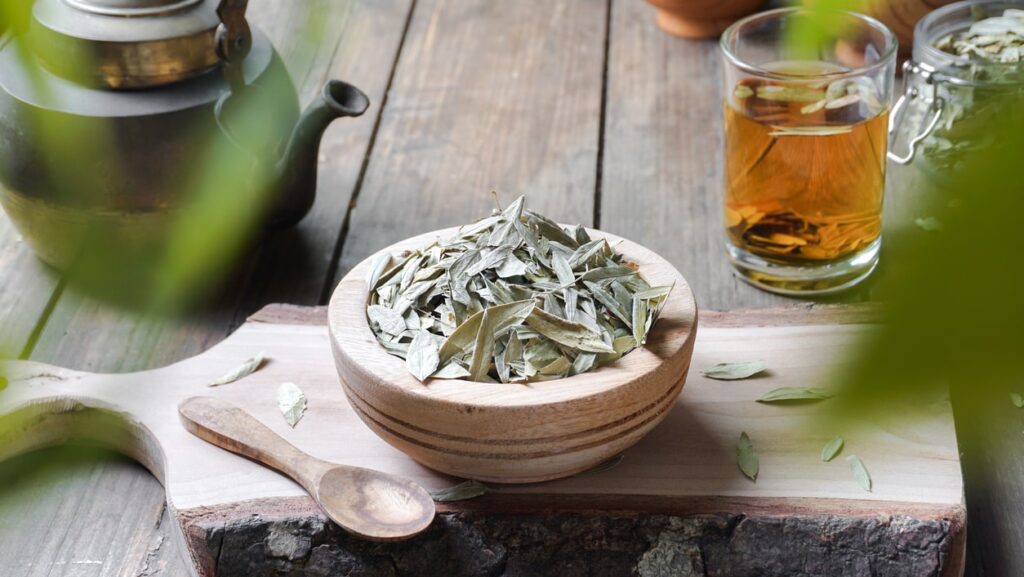 Wooden bowl of dried senna leaves beside a glass of senna tea and a teapot